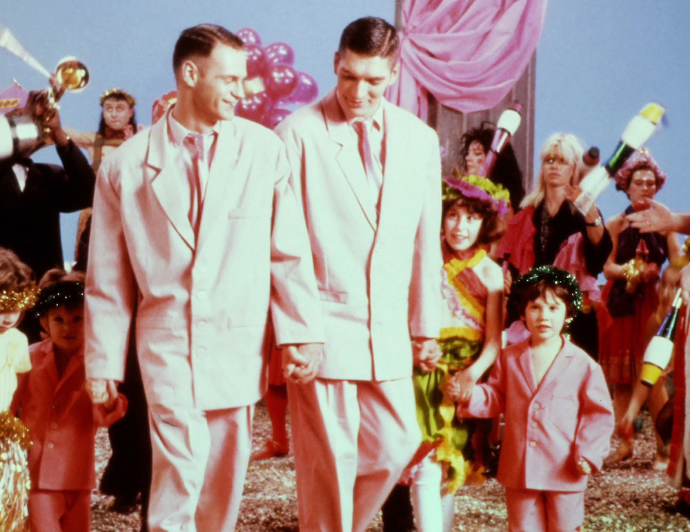 A still from Derek Jarman&rsquo;s &lsquo;The Garden&rsquo;. Two people in pink suits hold hands, walking, surrounded by ballons, decorations, people celebrating and making music.